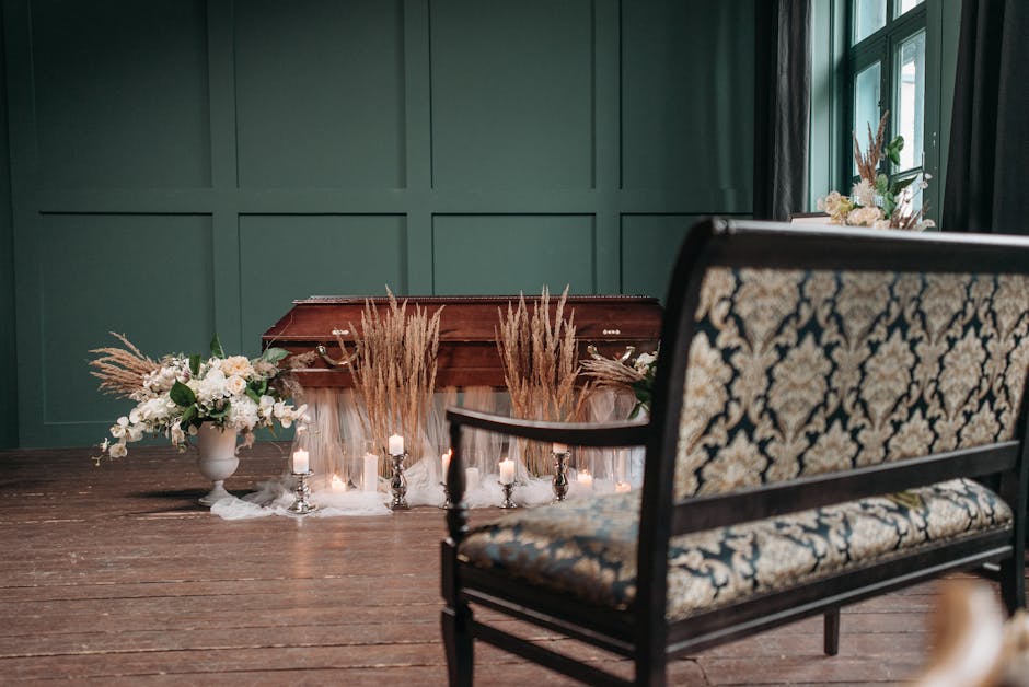 A flower arrangement situated on a wooden floor in front of a green paneled wall, featuring white and blush pink flowers in a white ceramic vase, complemented by dried pampas grass. Small white candle