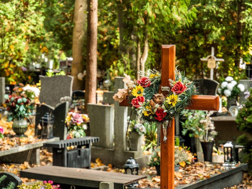 A wooden funeral cross adorned with a floral arrangement featuring red roses, yellow daisies, and pale pink flowers, surrounded by green foliage, situated in a graveyard with tombstones and memorial p