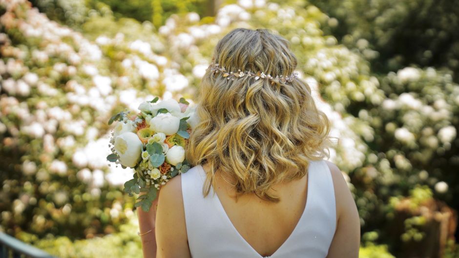 A bride with shoulder-length, curly blonde hair wearing a delicate floral headpiece stands outdoors, holding a floral bouquet featuring white peonies, soft pink roses, eucalyptus leaves, and other pas