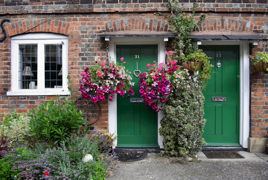 A charming brick building featuring two green front doors labeled with house numbers 31 and 33, each adorned with a brass letterbox and door knocker. Surrounding the doors are vibrant floral displays,