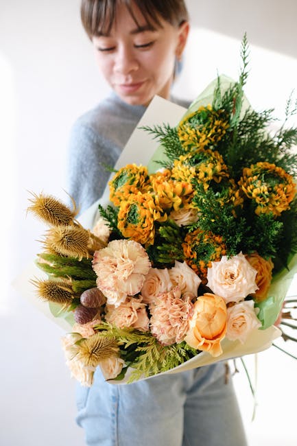 A woman holding a large floral bouquet featuring a mix of fresh flowers, including soft peach and cream roses, vibrant yellow and orange craspedia flowers, and green foliage, wrapped in pastel green p