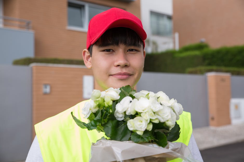 A young man with fair skin wearing a red cap and a high-visibility yellow vest is smiling and holding a bouquet of fresh white flowers, including roses and freesias, wrapped in light brown paper. He i
