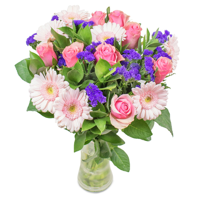 A vibrant and artfully arranged bouquet, set in a crystal-clear glass vase, is displayed against a pristine white backdrop-an image that could easily grace a window near Tunbridge Wells' Pantiles. The central focus is an exquisite collection of about seven to nine soft and medium pink roses, their elegant spiral petals unfurling in shades from pale blush to warm rosewood. At their side, five to seven pale pink gerbera daisies present large, welcoming faces, each with delicately textured, radiating petals and creamy, near-white centres. Threaded throughout are clusters of brilliant purple statice flowers, whose papery texture and rich colour add both energy and contrast to the otherwise gentle pink palette. Lush emerald-green foliage surrounds the blossoms, adding visual depth, natural movement, and the quiet suggestion of abundant health and vitality commonly seen in the green spaces around Dunorlan Park. The transparent glass vase highlights the neatly trimmed stems and clear water, reinforcing the impression of a just-delivered local bouquet. This refined arrangement could celebrate a special birthday, an anniversary, Mother's Day, or simply bring colour and freshness into a Tunbridge Wells office or home. Each stem is chosen and arranged with a florist's practiced hand, so the completed bouquet radiates a sentiment of affection, joy, and celebration deeply rooted in the character of Tunbridge Wells.